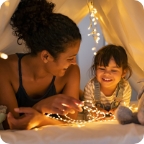 Mother and young daughter sit in a living room fort made of sheets.